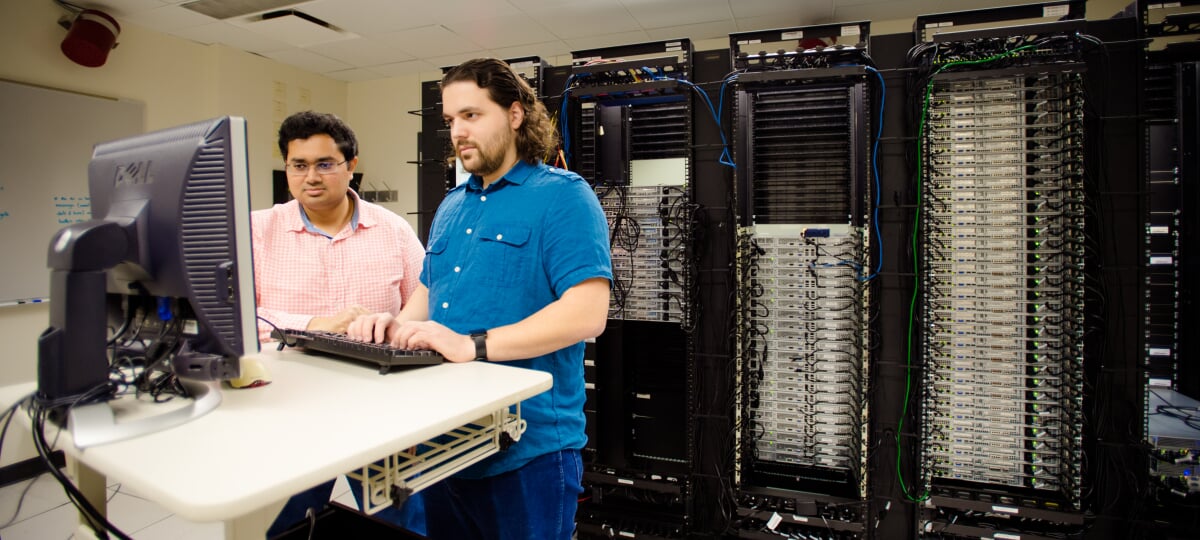 Two graduate students working at a standing desk with a monitor and keyboard, in front of a row of server racks.