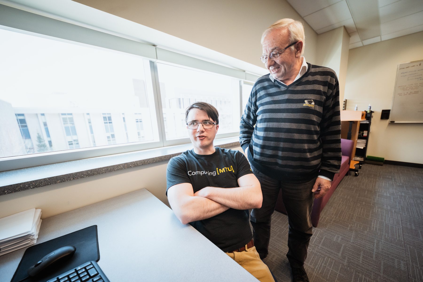 Prof. Önder standing beside a seated student, in conversation next to a window.