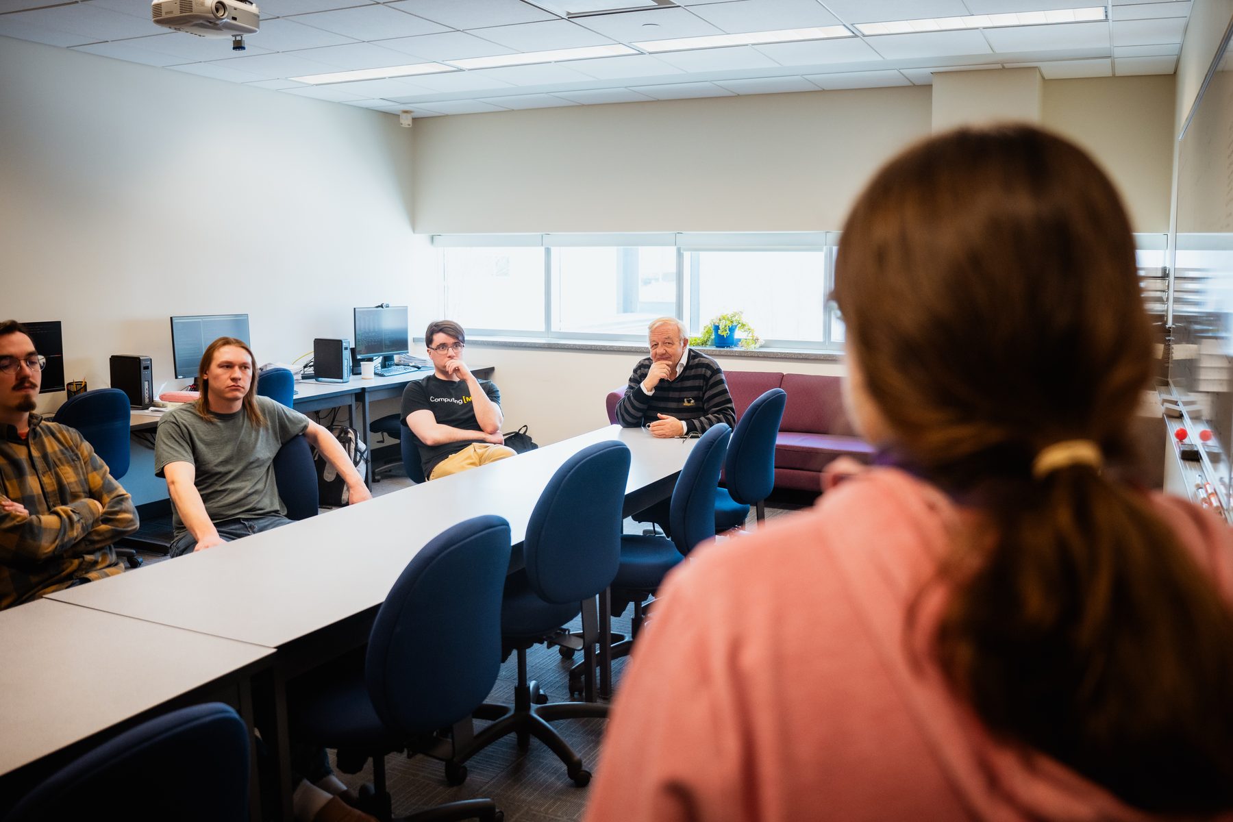 A student presents at a whiteboard while the rest of the group listens from around the table.