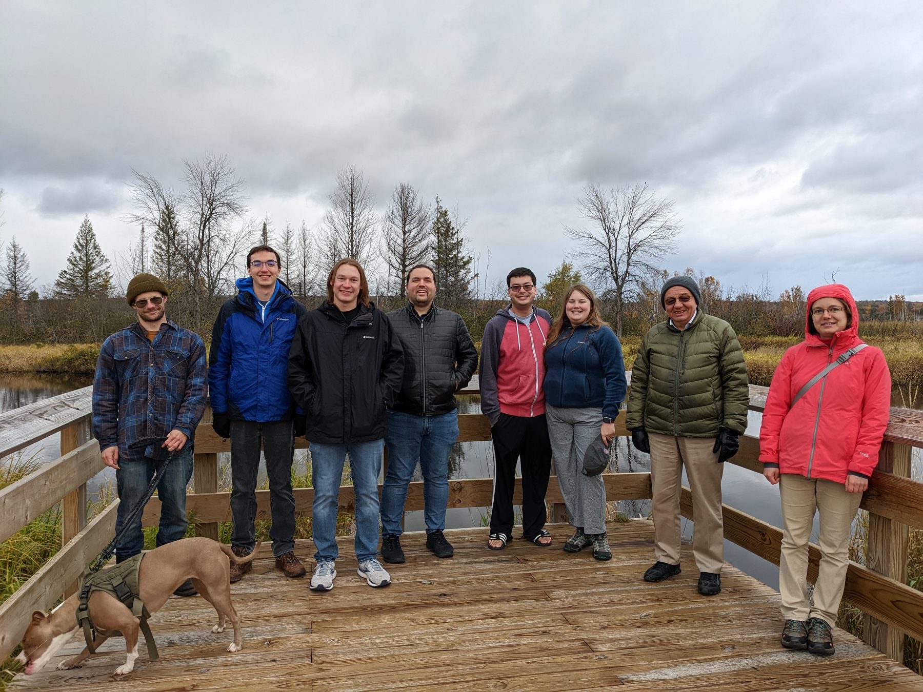 Eight lab members and a dog on a wooden boardwalk at Nara Nature Park, with marsh and bare trees behind them.