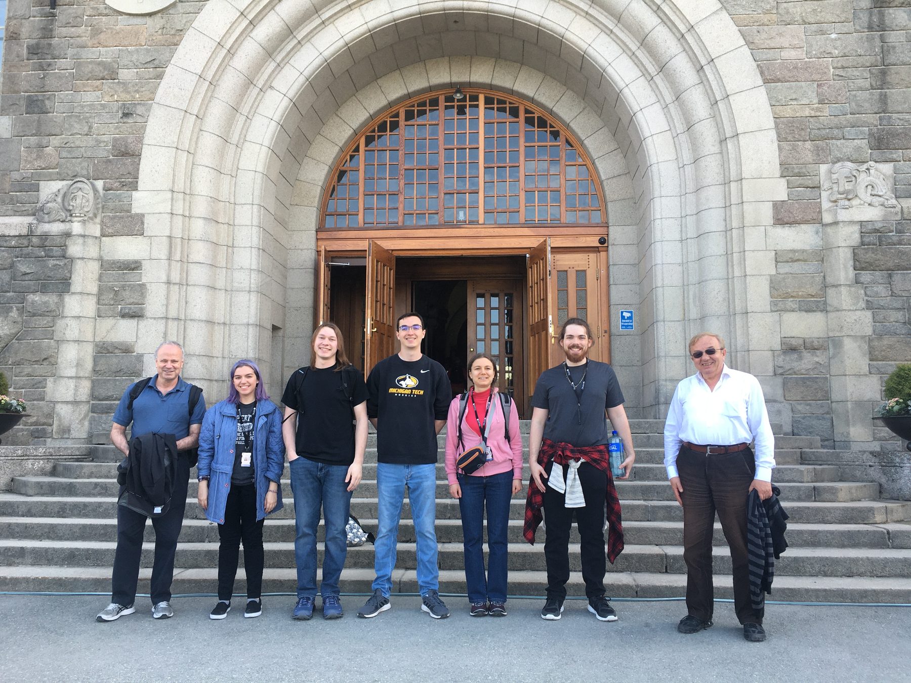 Prof. David Whalley, five students, and Prof. Soner Önder standing on the stone steps of NTNU's main building in Trondheim, in front of a large arched wooden doorway.